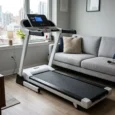 Tapis de marche pliable inclinable installé dans un salon près d’un canapé, avec vue urbaine par la fenêtre.